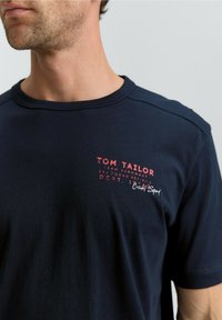 Navy Baumwoll-T-Shirt mit Rundhalsausschnitt, kurzen Ärmeln und weiß/rot bedrucktem Text auf der Brust, mit einem glatten, leicht strukturierten Finish.