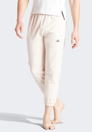 Homme marchant pieds nus, portant un jogging Adidas beige clair et une chemise blanche, visible de la taille aux pieds sur un fond uni.