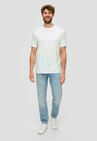 Biała bawełniana koszulka z zielonymi haftowanymi wzorami w gwiazdki, zestawiona z jasnoniebieskimi dżinsami i białymi trampkami. Model stoi i się uśmiecha.