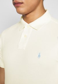 Žluté polo tričko z texturované látky, s klasickým límečkem a dvěma knoflíky, s malým modrým vyšívaným logem na hrudi.