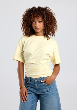Woman with curly hair wearing a pale yellow short-sleeve top and blue jeans, posing with one hand in pocket and neutral expression.