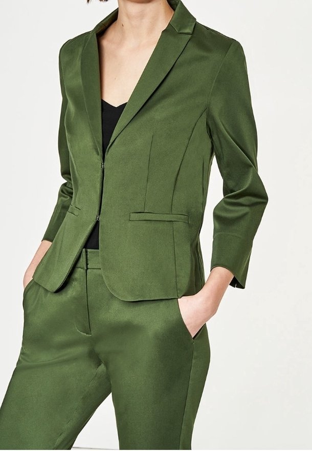 Femme portant un blazer olive vert ajusté et un pantalon assorti, les mains dans les poches, sur un haut noir, devant un fond uni.