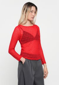 Rød blonde langermet topp med et semi-transparent design, rund halsudskæring og tettsittende overdel. Kombinert med høy midje grå bukser.