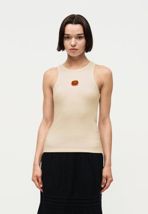 Frau mit kurzen dunklen Haaren, die ein beige ärmelloses Oberteil mit einem orangefarbenen Krabbenmotiv und einem schwarzen Rock vor einfachem Hintergrund trägt.