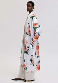 Lång blommig kimono i vitt med stora rosa och gröna blommor. Har krage, vida ärmar och en fransad fåll. Bärs över beige byxor.