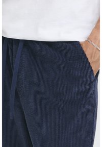 Námořnické kordurové tepláky s vertikální žebrovanou strukturou, šňůrkou v pase a bočními kapsami, částečně viditelné pod bílou košilí.