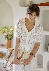 Femme souriante regardant vers le bas, portant un haut blanc à maille ouverte boutonné, tenant un sac en paille tissé avec des anses en cuir à l'intérieur.