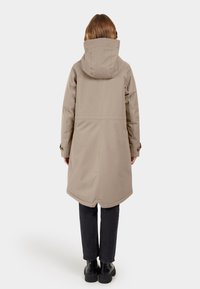 Beige lange jas met een capuchon, met een gladde, matte textuur en een rechte snit. Draagt over zwarte broek en stevige zwarte laarzen.