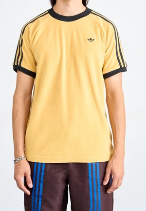 Person trägt ein gelbes Adidas-T-Shirt mit schwarzen Streifen an den Ärmeln und einem kleinen Logo, kombiniert mit dunklen Shorts mit blauen Streifen.
