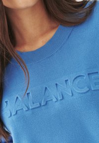Plavi pulover s rebrastim izrezom i teksturiranim natpisom "BALANCE" sprijeda; mekan trikotažni materijal i glatka tekstura.