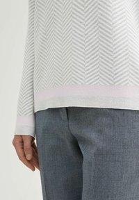 Grijs en wit gebreid sweater met een visgraatdesign en een roze accent aan de zoom, gecombineerd met grijze broek. Close-up van het onderste deel van de outfit.