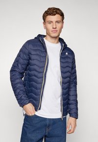 Manteau rembourré bleu marine avec un matelassage en chevrons, fermeture éclair à l'avant et capuche. Présente un petit logo sur la poitrine et des poignets élastiques.
