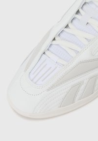 Sneaker alb cu un upper din piele texturată, accente gri deschis, limba căptușită și șireturi plate. Prezintă cusături distincte și deschideri de ventilație.