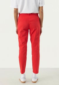 Rote, schlank geschnittene Hose aus leichtem Material mit mittelhoher Taille, zwei Gesäßtaschen und schmal zulaufendem Bein. Getragen mit weißen Schuhen.