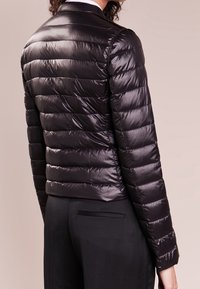 Chaqueta acolchada negra con un acabado brillante, que presenta un diseño corto, costuras horizontales y un cuello clásico. Se lleva con pantalones negros.