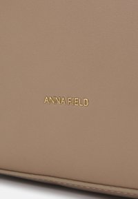 Beige keinomateriaali sileällä pinnalla. Kultaprintattu "ANNA FIELD" -logo. Minimalistinen muotoilu hienovaraisilla ommellaan. 