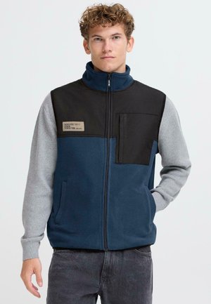 Veste en polaire avec un corps bleu marine, une partie supérieure noire et des manches grises. Elle possède une fermeture éclair et une poche poitrine avec une étiquette.