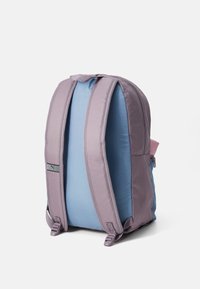 Mochila de tela en púrpura claro y azul, con correas de hombro acolchadas, un bolsillo lateral con cremallera y un pequeño logotipo como acento.