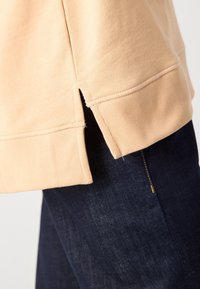 Ourlet de sweat beige avec fente latérale sur jean en denim bleu foncé, détail en gros plan.