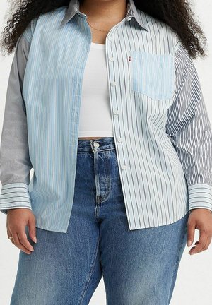 Osoba ubrana w zapinaną koszulę z mieszanym wzorem pasków, na białym topie odsłaniającym brzuch, w połączeniu z wysokim stanem niebieskich dżinsów.