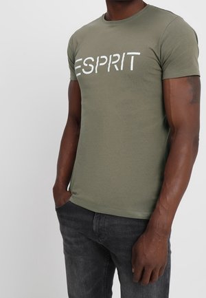 Man draagt een olijfgroen T-shirt met korte mouwen met het "ESPRIT" logo en donkergrijze jeans, staand met één hand in de zak.