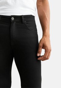 Svarta denimjeans med slim fit. Funktioner inkluderar en metallknapp, bältesöglor och en slät textur. Handen är placerad vid midjan.