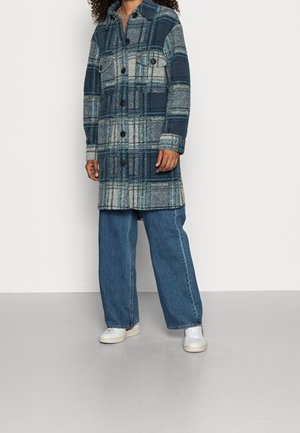 Persona con un abrigo abotonado a cuadros azul y gris, vaqueros anchos azules y zapatillas blancas de pie frente a un fondo liso.
