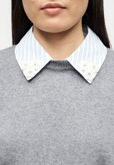 Gros plan d'une personne portant un pull gris par-dessus une chemise à col rayée blanche et bleue ornée de motifs floraux blancs sur le col.