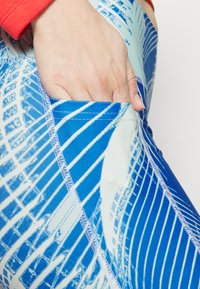 Leggings blu con motivo presentano un design astratto con sfumature di blu e bianco. Una mano sta infilando qualcosa in una tasca laterale. Dettagli delle cuciture visibili.