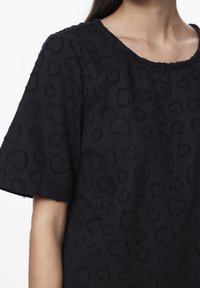 Black short-sleeve top with textured heart patterns worn by a person, visible from shoulder to mid-upper arm and neck.