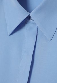 Cămașă albastră deschis cu guler punctat, având închidere cu buton și țesătură texturată. Cusăturile sunt vizibile de-a lungul plăcătorului.