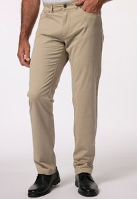 Beżowe spodnie slim fit wykonane z bawełny, z zapięciem na zamek błyskawiczny i guzik, z bocznymi kieszeniami i nogawkami zwężającymi się ku kostce, noszone z czarnymi butami.
