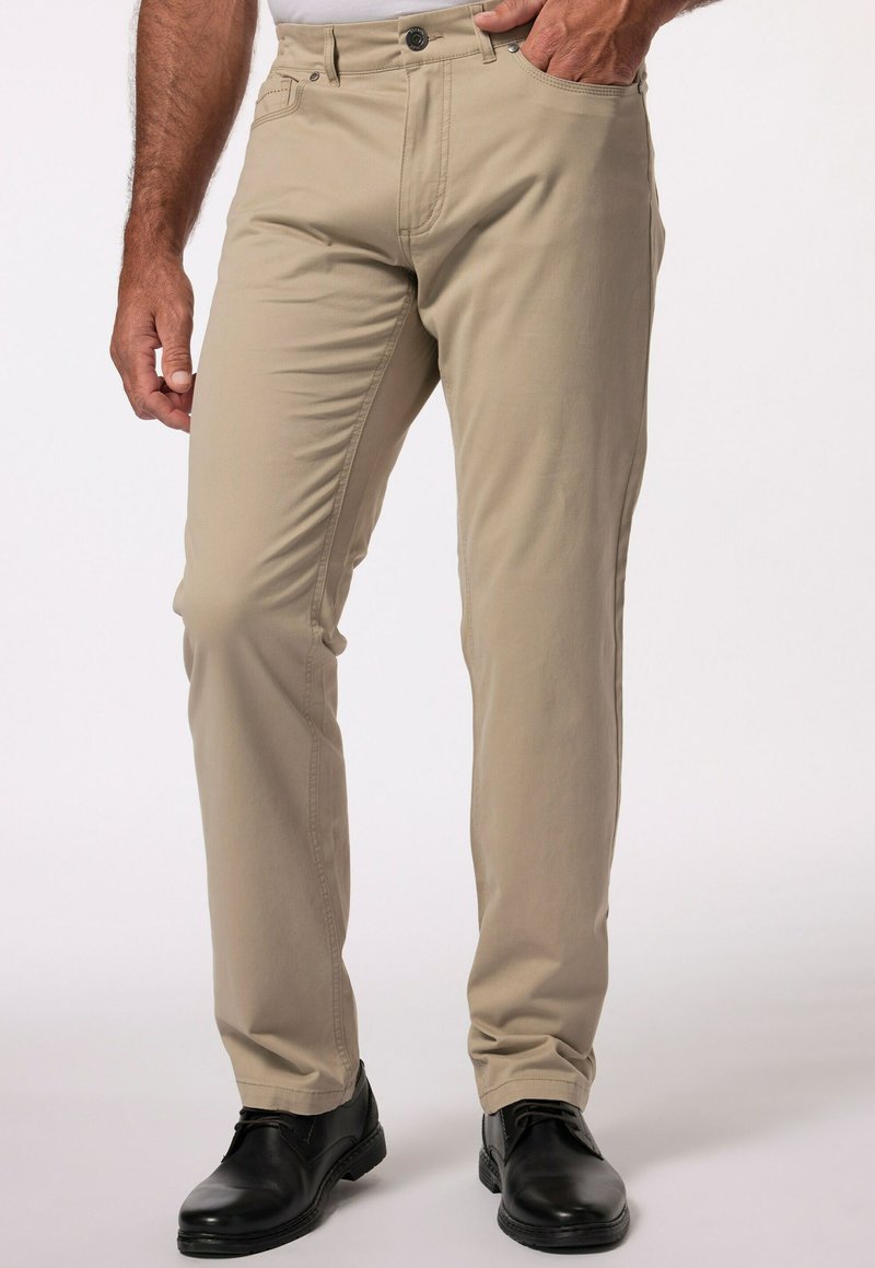 Beżowe spodnie slim fit wykonane z bawełny, z zapięciem na zamek błyskawiczny i guzik, z bocznymi kieszeniami i nogawkami zwężającymi się ku kostce, noszone z czarnymi butami.