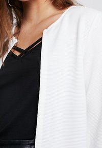 Cardigan bianco a coste realizzato in materiale testurizzato, design a apertura frontale, indossato sopra un top nero a strati con spalline incrociate.