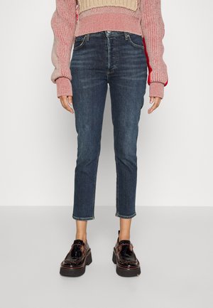 Person iført cropped mørkeblå jeans, lyserød strikket sweater og sorte skinnende platåsko, der står på en neutral baggrund.