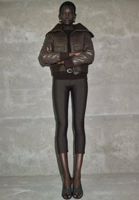 Giubbotto in pelle marrone cropped con design a trapunta, top aderente nero, leggings scuri e mules aperti neri, posato su uno sfondo neutro.