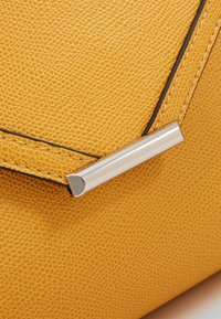 Žlutá texturovaná kožená kabelka, roh s viditelným stříbrným kovovým uzávěrem a černým lemováním.