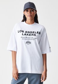 Bijela oversized majica s natpisom "LOS ANGELES LAKERS" u crnom podebljanom tekstu, uz dodatno brendiranje. Nosiva s sivom kapom, ležerna tkanina.