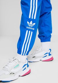 Pantalones deportivos azules con diseño de tres rayas blancas y logo de Adidas, combinados con zapatillas blancas y rosas con acentos azules y superficies texturizadas.