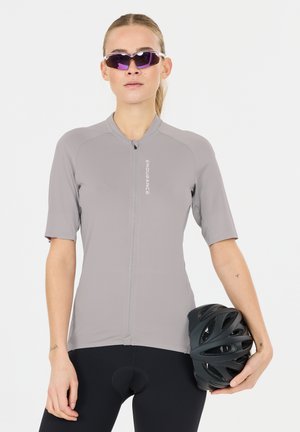 Femme portant un maillot de cyclisme gris et des lunettes de soleil, tenant un casque de vélo noir à côté d'elle.