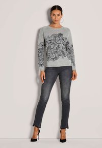 Frau, die einen grauen Pullover mit schwarzem floralem Paisleymuster und dunklen grauen Skinny-Jeans trägt, steht vor einem neutralen Hintergrund.