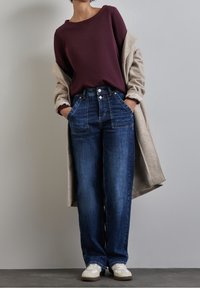 Sudadera color burdeos, cárdigan largo beige, jeans azules de tiro alto con bolsillos frontales y zapatillas blancas, sobre un fondo neutro.