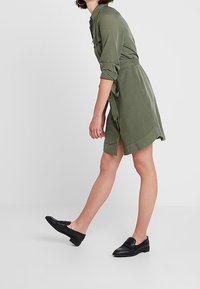 Olivově zelené šaty s dlouhými rukávy, s páskem na uvázání a bočními rozparky. Spárováno s černými nazouvacími botami, které mají plochou podrážku a hladký povrch.