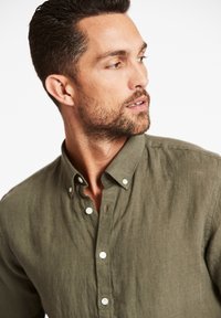 Olijfgroene button-up shirt met een zachte textuur, voorzien van een klassieke kraag en witte knopen, gedeeltelijk zichtbaar op de bovenlichaam van een model.