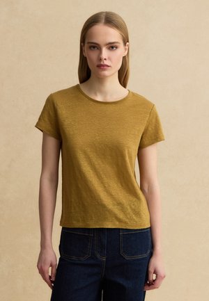 Giovane donna con capelli biondi lisci che indossa una maglietta giallo senape a maniche corte e jeans blu scuro, in piedi davanti a uno sfondo beige uniforme.