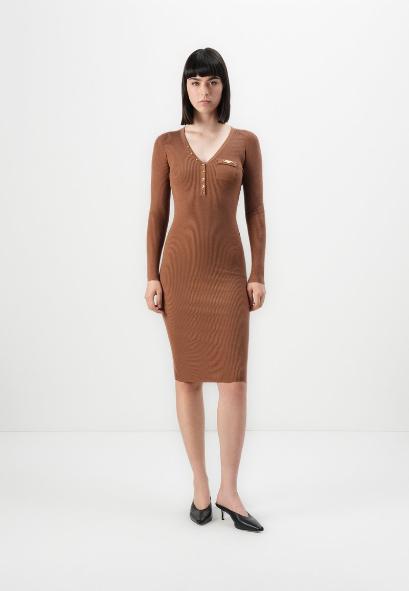 Vestido de malha canelada castanho com mangas longas, decote em V profundo, detalhe em botão dourado no peito e uma silhueta justa, combinado com mules pretas de salto.