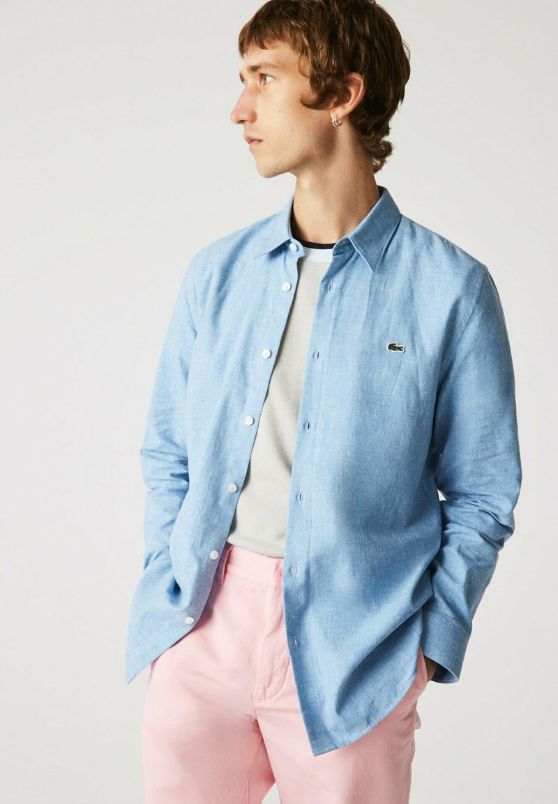 Helles blaues Hemd mit Struktur aus gewebtem Stoff, mit Kragen und einem kleinen grünen Logo. Kombiniert mit hellroten Hosen.