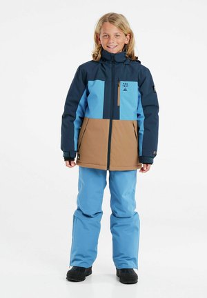 Giacca da neve blu e marrone con zip frontale, cappuccio e design a blocchi di colore. Pantaloni blu abbinati e stivali invernali neri.