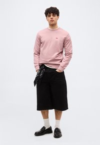 Νεαρός με σγουρά μαλλιά που φοράει ροζ ανοιχτό φούτερ, μαύρη βερμούδα μέχρι το γόνατο με μαύρο μαντίλι, λευκές κάλτσες και μαύρα παπούτσια.