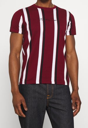 Homme portant un t-shirt bordeaux à manches courtes avec des rayures verticales blanches et noires et un jean bleu foncé, debout avec les bras détendus.